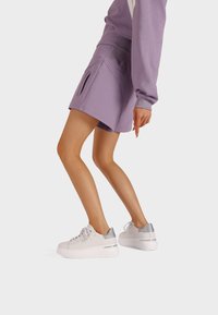 Pantaloni sportivi viola con tasche laterali. Abbinati a sneakers bianche con un accento metallico argento sul tallone e una suola spessa bianca.