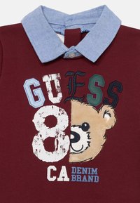 Chemise bordeaux avec un col en denim bleu clair. Présente un graphisme d'ours, des chiffres surdimensionnés "8" et le texte "GUESS CA DENIM BRAND" dans différentes couleurs.
