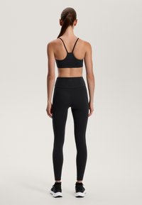 Musta urheiluliivi, jossa on ohuet hihat, yhdistettynä korkeavyötäröisiin mustiin legginsseihin. Sujuva, joustava kangas. Malli kääntää selkänsä, päällään mustat lenkkarit.