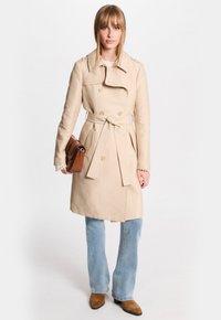 Beige trenchcoat met dubbele knopen, een ceintuur op de taille en schouderepauletten. Draag het over lichtblauwe flared jeans en bruine laarzen.