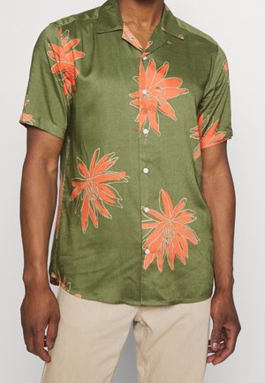 Man draagt een olijfgroen overhemd met korte mouwen met grote oranje bloemenprints en een beige broek tegen een effen achtergrond.