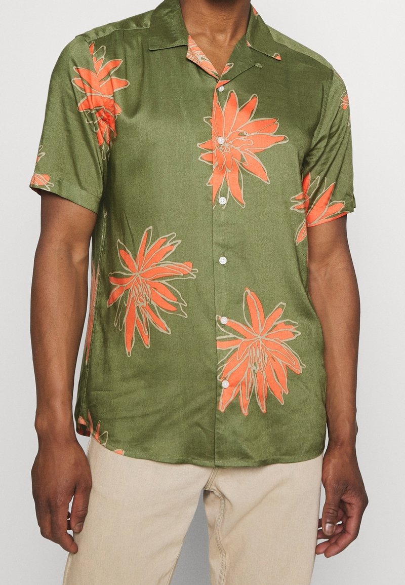 Homme portant une chemise boutonnée olive à manches courtes avec de grandes imprimés floraux orange et un pantalon beige sur un fond uni.