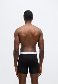 Chiloți boxeri negri cu o talie albă pe care este inscripționat logo-ul „Calvin Klein”, priviți din spate, evidențiind materialul neted și croiala.