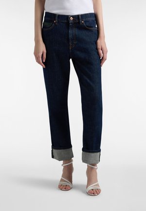 Denim jeans in dark wash with a relaxed fit, rolled cuffs, and orange stitching details. Paired with white strappy sandals.