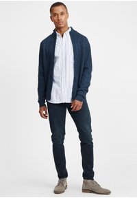 Sötétkék kötött kardigán texturált mintával, fehér és kék csíkos ing fölött viselve. Sötét farmernadrággal és világosszürke velúrcipővel párosítva.