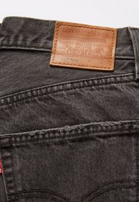 Vaqueros de mezclilla gris oscuro con un parche de cuero con marca en relieve y detalles de costura, con una pestaña roja en el lado izquierdo.