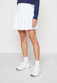 Bílá plisovaná sukně s hladkým povrchem a elastickým pasem, spojená s bílými teniskami a kotníkovými ponožkami, pohled z pravé strany.