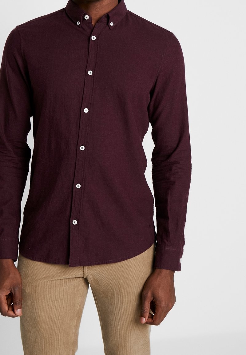 Chemise bordeaux à manches longues avec col boutonné, dotée de boutons blancs, d'une texture fine et d'un ourlet arrondi. Associée à un pantalon beige clair.