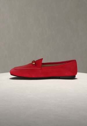 Mocassin plat en daim rouge avec un détail boucle dorée sur le dessus, présenté sur un fond gris neutre et une surface claire.