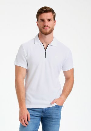 Camisa polo blanca de manga corta con un patrón texturizado, cierre de cremallera y un pequeño logo en el pecho, combinada con jeans de un azul claro.