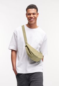 Joven sonriendo, vistiendo una camiseta blanca, jeans grises y un bolso cruzado de color oliva claro frente a un fondo blanco uniforme.