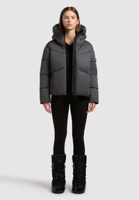 Grå polstret jakke med hætte, lynlås foran og elastiske detaljer. Kombineret med tætsiddende sorte bukser og blanke sorte støvler.