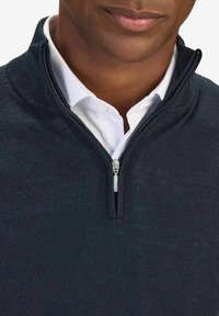 Mörk marinblå zip-up tröja med en strukturerad stickad yta, hög krage och silverfärgad dragkedja, lagd över en vit skjorta med krage.