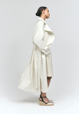 Femme avec les cheveux en chignon portant un long trench-coat écru et des espadrilles compensées beiges avec des brides noires à la cheville, debout de profil.