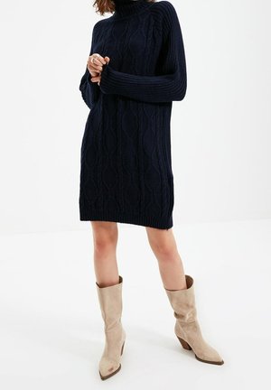 Femme portant une robe-pull en maille torsadée bleu marine à manches longues et des bottes mi-mollet en daim beige, debout sur fond blanc.