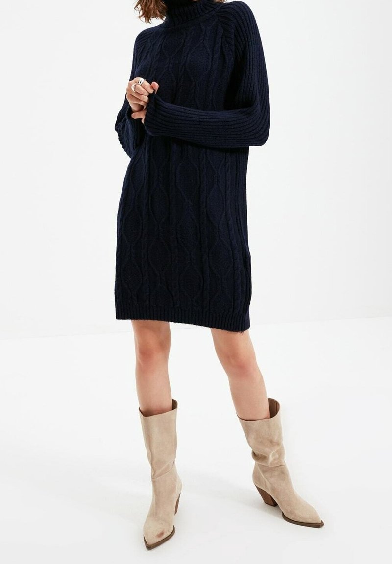 Femme portant une robe-pull en maille torsadée bleu marine à manches longues et des bottes mi-mollet en daim beige, debout sur fond blanc.