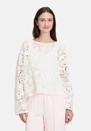 Top en crochet blanc avec motif floral, manches larges et coupe décontractée. Porté sur un pantalon rose clair pour un look en superposition.