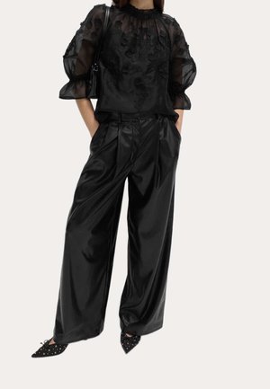 Mujer con blusa negra transparente con flores y mangas abullonadas, pantalones negros de satén de talle alto y holgados, zapatillas negras puntiagudas con detalles de ojales y un bolso de hombro.