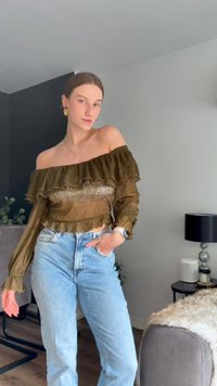 Top verde-oliva com decote à off-the-shoulder, com franzidos e detalhes em renda, combinado com jeans de cintura alta em azul claro. Estilo casual moderno.