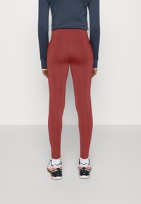 Rode legging met een hoge tailleband, gladde textuur en gestikte achterzakken, gecombineerd met zwart-oranje sportschoenen.