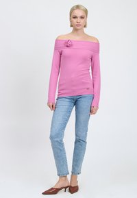 Rosa schulterfreier Pullover mit Rosendetail, taillierte Passform, gerippte Bündchen, kombiniert mit hellblauen Jeans und braunen spitzen Mules.