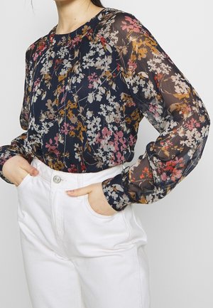 Blumenhafte transparente Bluse in Marineblau mit langen Ärmeln, rundem Halsausschnitt und bunten Blumenmustern. Kombiniert mit hoch taillierten weißen Hosen.