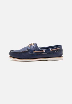 Chaussure bateau en cuir bleu marine avec lacets en cuir couleur noisette et semelle blanche, vue de côté sur fond blanc.