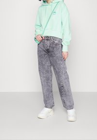 Miętowo-zielona, skrócona bluza z kapturem z logo, w zestawieniu z wysokimi, prostymi, wyblakłymi czarnymi dżinsami oraz białymi trampkami o teksturowanej podeszwie.