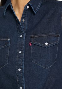 Chemise en denim bleu foncé avec poches à rabat boutonnées, boutons-pression blancs et surpiqûres beiges visibles sur le col et la patte de devant.