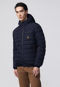 Uomo che indossa una giacca imbottita con cappuccio di colore blu navy, dotata di tasche frontali, insieme a pantaloni marroni, in piedi contro uno sfondo chiaro e semplice.