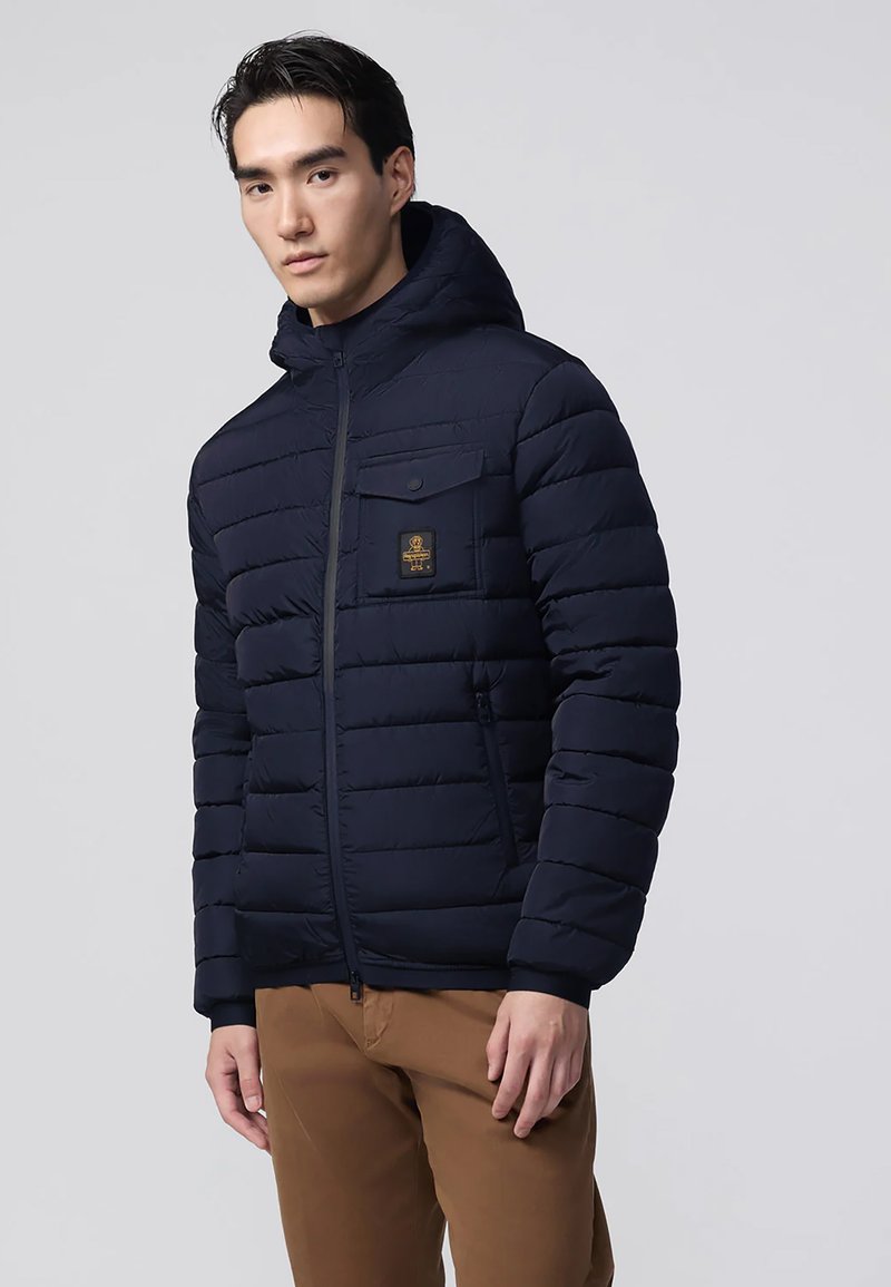 Uomo che indossa una giacca imbottita con cappuccio di colore blu navy, dotata di tasche frontali, insieme a pantaloni marroni, in piedi contro uno sfondo chiaro e semplice.