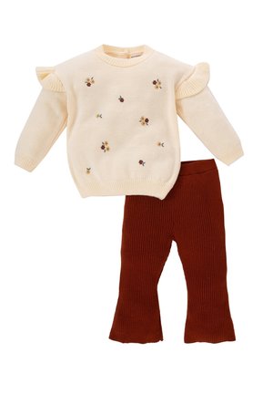 Cremefarbener Strickpullover mit kleiner Blumenstickerei und gerüschten Schultern, kombiniert mit rostfarbenen, gerippten Strickhosen.