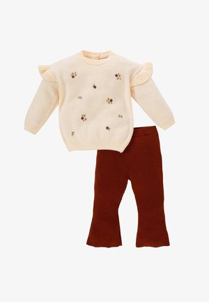 Kremowy sweter z dzianiny z małą kwiatową haftem i falbanami na ramionach, zestawiony z spodnimi z dzianiny prążkowanej w kolorze rdzy.