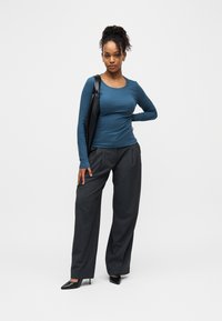 Γυναίκα με καστανά σγουρά μαλλιά, φορώντας μπλε μπλούζα με μακριά μανίκια και γκρι παντελόνι με φαρδύ πόδι, στέκεται με μαύρη τσάντα ώμου και μαύρες ψηλοτάκουνες γόβες.