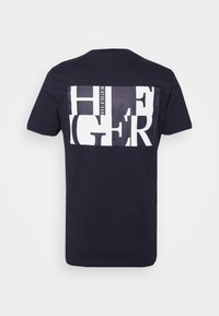 Námorně modré bavlněné tričko s velkým bílým grafickým designem na zadní straně, který obsahuje bloková písmena a název značky v kontrastních stylech.