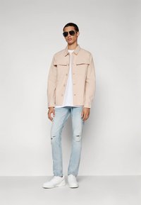 Casaco bege claro com botões e dois bolsos no peito, usado por cima de uma t-shirt branca; combinado com jeans azuis claros rasgados e ténis brancos.