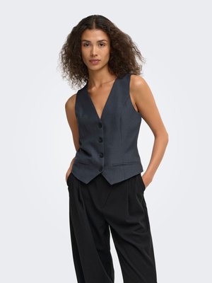 Femme aux cheveux bouclés portant un gilet gris foncé boutonné et un pantalon noir ample, debout les mains dans les poches sur un fond clair.