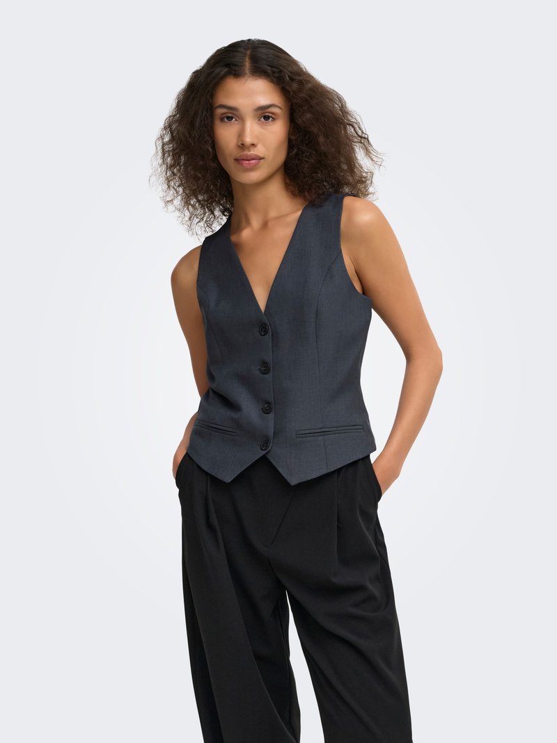 Femme aux cheveux bouclés portant un gilet gris foncé boutonné et un pantalon noir ample, debout les mains dans les poches sur un fond clair.