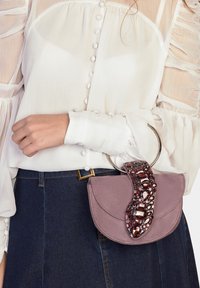 Borsa in pelle scamosciata viola con una forma curva, dotata di una tracolla di perline e di un manico circolare in metallo. La borsa è parzialmente appoggiata a una camicetta bianca.