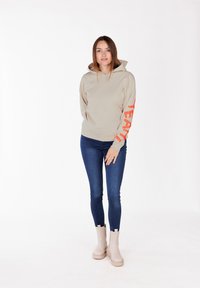 Felpa beige con cappuccio e testo arancione sulla manica sinistra, abbinata a jeans blu attillati e stivali alla caviglia beige. Texture del tessuto liscia.