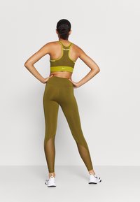 Oliwkowy stanik sportowy z żółtymi akcentami, w połączeniu z legginsami o wysokim stanie z siateczkowymi wstawkami i gładką teksturą. Białe sneakersy w zestawie.