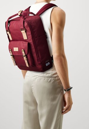 Personne portant un haut blanc sans manches et un pantalon beige avec un sac à dos bordeaux fixé par des sangles en cuir dans le dos.