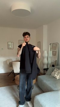 Sweater preto segurado por uma pessoa, com um corte casual e mangas compridas. O ambiente contém móveis de cores claras e elementos de decoração neutros.