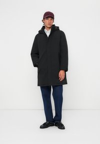 Zwarte parka met capuchon, gemaakt van waterbestendig materiaal. Voorzien van een rits aan de voorkant, zijzakken en een losse pasvorm. Gekocht met blauwe jeans.