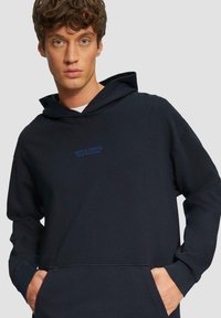 Donkerblauwe hoodie gemaakt van zacht stof, met een buidelzak aan de voorkant, geribbelde mouwen en een "Arts & Crafts" logo in lichtblauw op de borst.