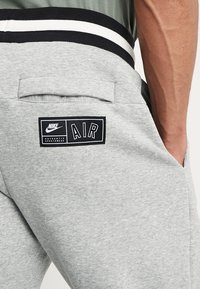 Šedé tepláky Nike s černobílým páskem v pase, s tištěnou nášivkou loga "AIR" na zadní kapse.
