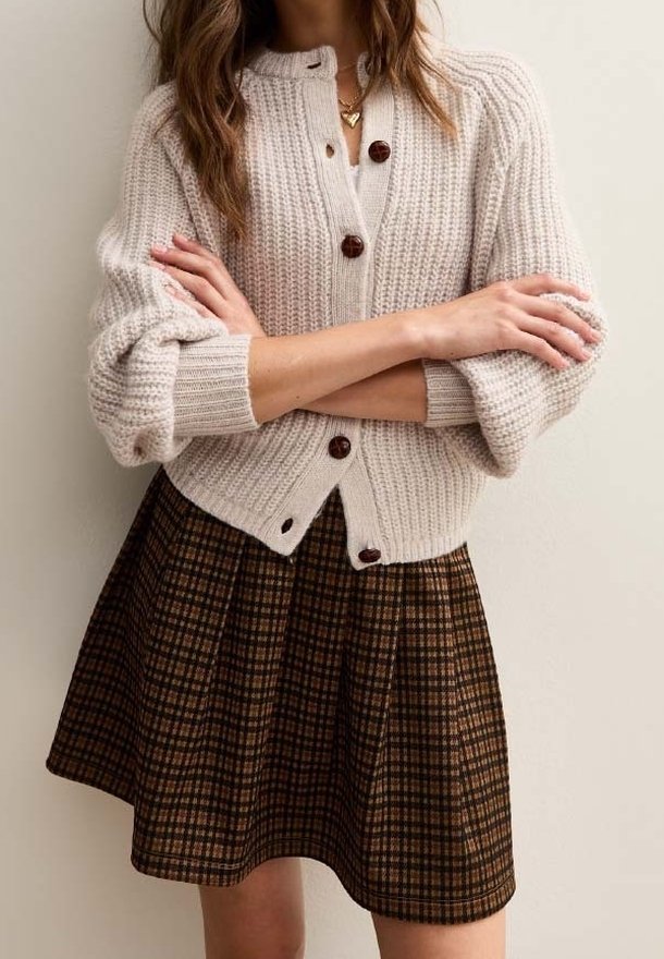 Femme portant un cardigan en grosse maille crème avec de gros boutons sur une jupe plissée à carreaux marron, bras croisés, debout devant un fond uni.