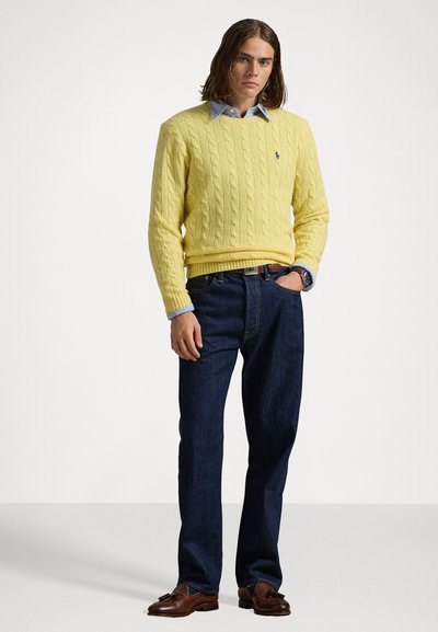 Dzeltena adīta džemperis, zem tā zila krekla apkakle, tumši džinsu bikses, brūni ādas apavi un pulkstenis uz rokas.