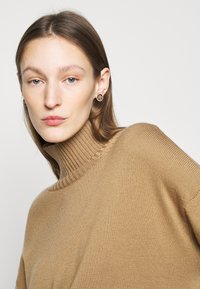 Bež polovično turtleneck pulover z ribastim ovratnikom in gladko teksturo, v kombinaciji z okroglimi, okrašenimi uhani. Portret v ozkem kadru.