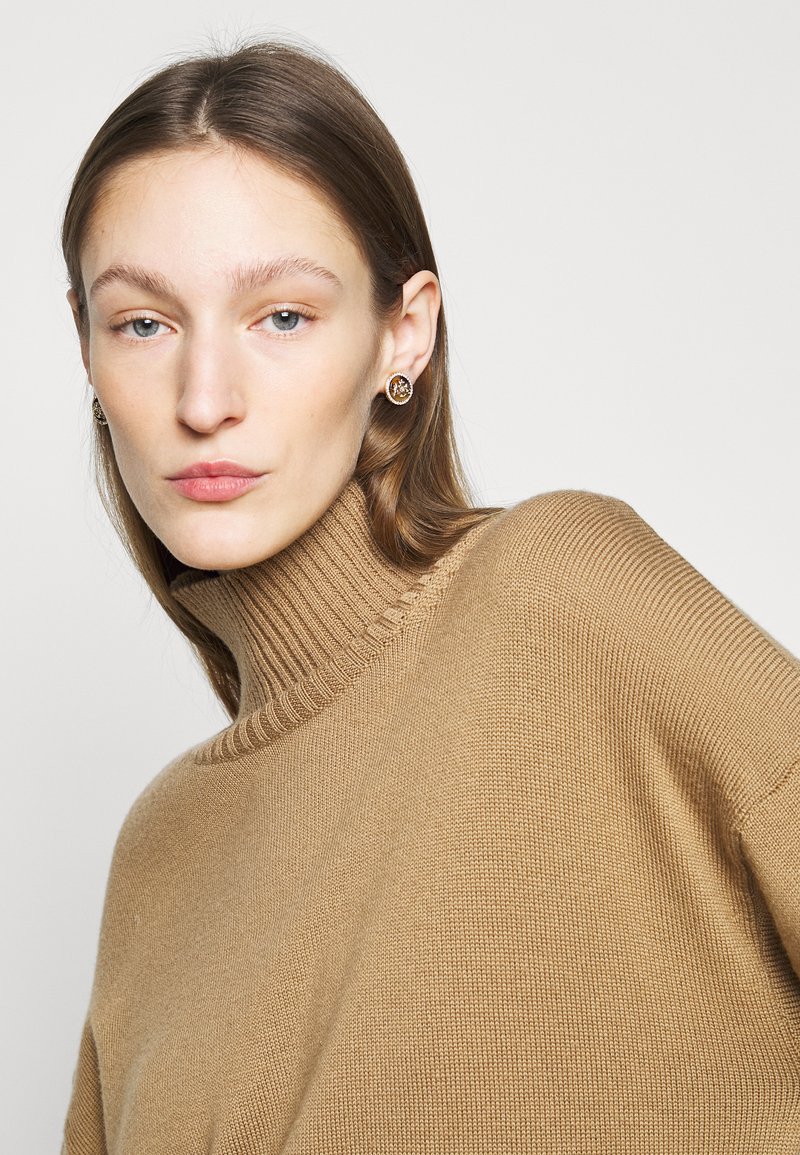 Bež polovično turtleneck pulover z ribastim ovratnikom in gladko teksturo, v kombinaciji z okroglimi, okrašenimi uhani. Portret v ozkem kadru.